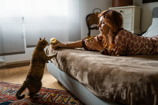 A woman lying on a bed lets her cat smell a jar of peanut butter
