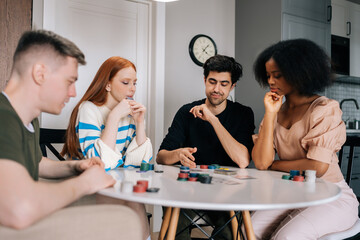 Portrait of multiethnic friends having home fun party on weekends playing poker game sitting at table in living room. Diverse young men and women satisfied with spending leisure together playing poker