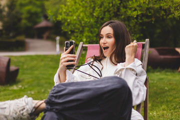 Happy smiling young woman hold smartphone with excited face expression, unable to believe that she bet played, win in lottery, read good news, can't believe in her fortune. Girl sitting in park.