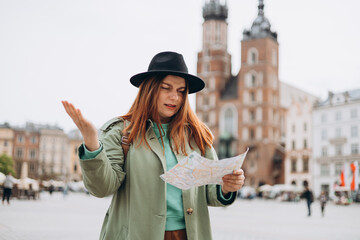 Sad young female tourist is lost in new city. Millennial Redhead girl walking in the park and holding a paper map and cannot find tourist destination in a sunny day in Krakow. St. Marys Basilica