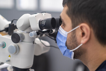 Closeup portrait of bearded dentist in medical mask on face looks in innovative dental microscope to examine teeth of patient professional services in contemporary clinic