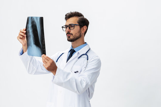 Male doctor in white coat and eyeglasses and stethoscope looks thoughtfully at pictures of X-ray patient for diagnosis on white isolated background, copy space, space for text, health