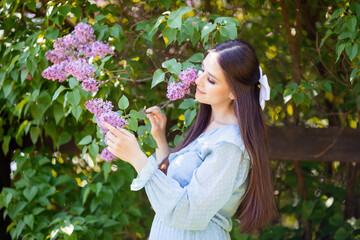  girl in blue dress stands nearby blooming lilac bush , in the garden