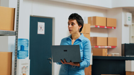 Female entrepreneur using laptop to do warehouse inventory, planning quality control for products before shipping them. Woman taking notes on pc and looking at merchandise packaging.