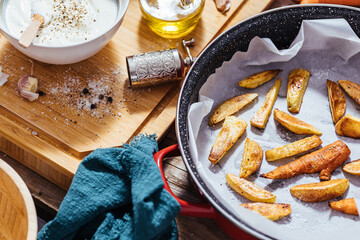 Baking Pan Filled with Roast Potatoes or Baked French Fries in Messy Working Kitchen with a Bowl of Yogurt Sauce, Olive Oil, Garlic and Spices and Condiments on Wooden Board and Table