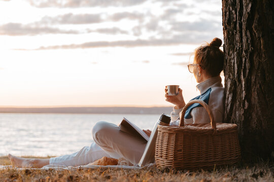 Woman drink tea or coffee from thermos read book having picnic at lake