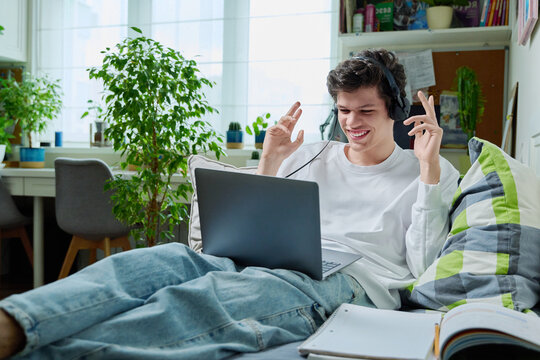 Young guy in headphones using laptop for video communication, lying at home