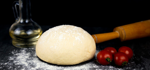 Cooking pizza, raw dough, rolling pin and cherry tomatoes on a black background. Pizza cooking concept.