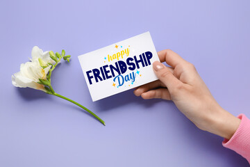 Female hand holding greeting card for Friendship Day on lilac background
