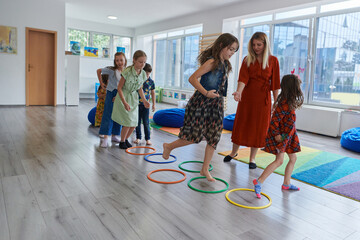 Obraz premium Small nursery school children with female teacher on floor indoors in classroom, doing exercise. Jumping over hula hoop circles track on the floor.