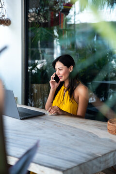 Smiling woman talking on smartphone working outdoors with laptop