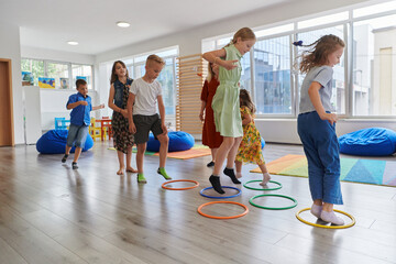 Small nursery school children with female teacher on floor indoors in classroom, doing exercise. Jumping over hula hoop circles track on the floor.