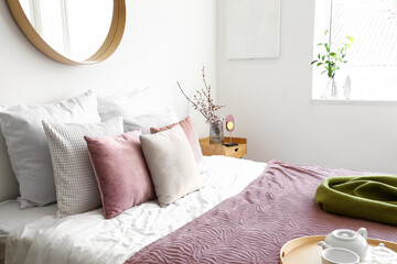 Cozy bed with many pillows and blooming tree branches on end table in bedroom