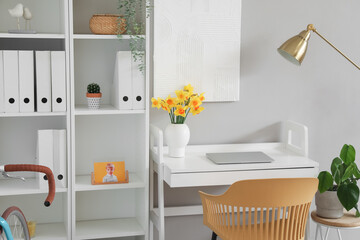 Comfortable workplace with modern laptop and narcissus flowers on table near grey wall