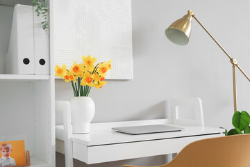 Comfortable workplace with modern laptop and narcissus flowers on table near grey wall