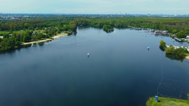Drone shot over Unterbacher See Lake in Dusseldorf, Germany. Beautiful blue fresh lake water and many green trees and bushes on the coast with beaches and boats sailing under the spring blue sky   