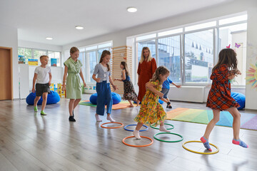 Small nursery school children with female teacher on floor indoors in classroom, doing exercise. Jumping over hula hoop circles track on the floor.