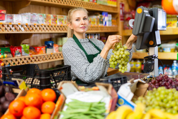 Focused saleswoman working at counter in local grocery store, weighing bunch of ripe grape on electronic scale..