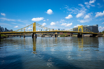 Obraz premium Iconic yellow Rachel Carson Bridge crossing the Allegheny River in Pittsburgh, Pennsylvania, with blue sky and white fluffy clouds.