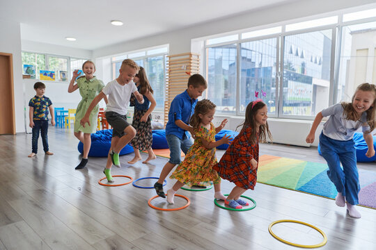 Small nursery school children with female teacher on floor indoors in classroom, doing exercise. Jumping over hula hoop circles track on the floor.