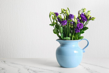 Jug with eustoma flowers on table near white wall