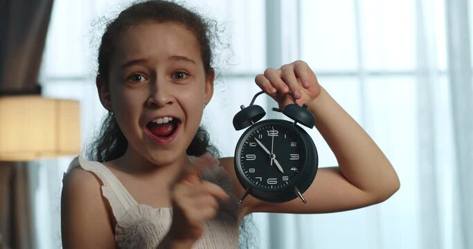 A happy cute little girl in a white dress holds an alarm clock in her hands, the alarm clock rings, the child makes an anxious and joyful face and says the time. Educational lifestyle. 4K