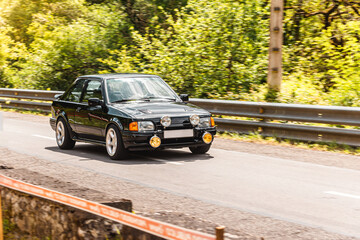 A classic rfetro european sport car is being driven fast by a competitor in a European hill climb championship in Asturias, Spain, in the middle of a forest and the mountains in a sunny daylight