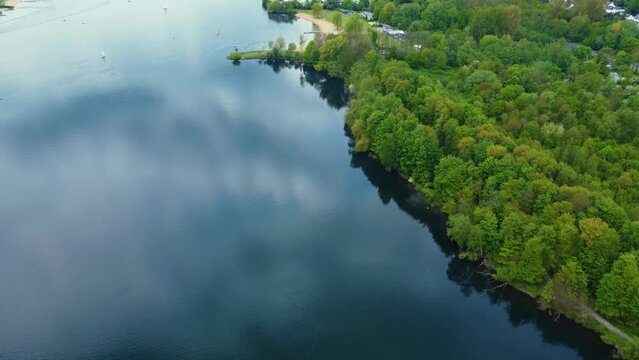 Drone shot over Unterbacher See Lake in Dusseldorf, Germany. Beautiful blue fresh lake water and many green trees and bushes on the coast with beaches and boats sailing under the spring blue sky   
