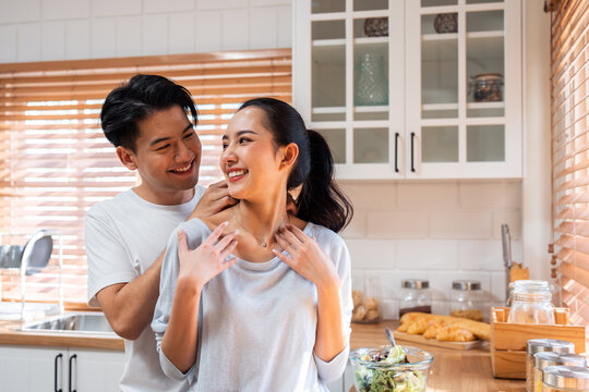 Asian romantic man making surprise girlfriend with necklace in kitchen. 