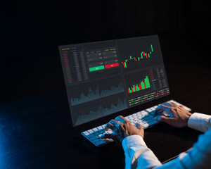 Caucasian man sits in the dark in front of a virtual screen. Stock exchange charts. Close-up of male hands on the keyboard.