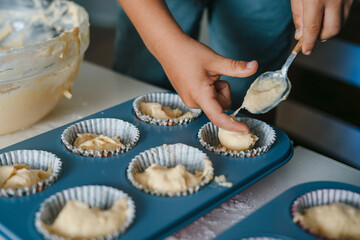 A child's hand pouring batter into cups during a home baking workshop, making cupcakes. Food preparation. Fun family.