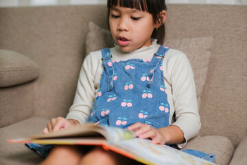 Naklejka premium Happy little girl reading a book indoors. Cute little girl in casual clothes sitting on the sofa reading a book.