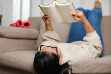 Fototapeta na wymiar Happy Asian woman lying on the sofa and reading a book indoors. Lifestyle, free time and relaxation.