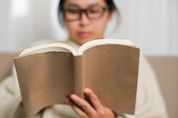 Happy Asian woman sitting on the sofa and reading a book indoors. Portrait of woman in white sweater reading a book on sofa at home. Lifestyle, free time and relaxation.