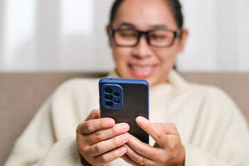 Happy young woman using mobile phone while sitting on sofa at home. Attractive woman holding smartphone checking social media, playing games, online shopping relaxing on sofa.