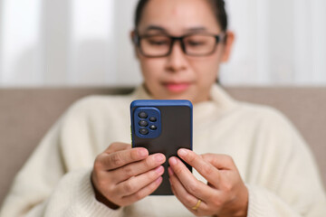 Happy young woman using mobile phone while sitting on sofa at home. Attractive woman holding smartphone checking social media, playing games, online shopping relaxing on sofa.