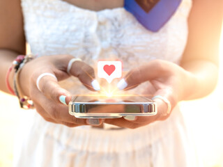 Giving heart, like symbol in speech bubble icon on social media on smart mobile phone in woman's hands using application. Close-up cell using by stylish female in white dress at the beach, summertime.