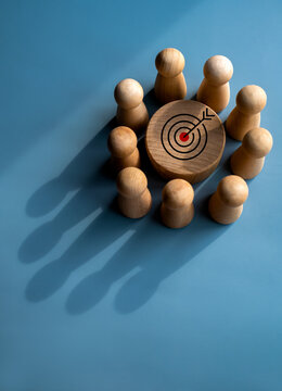 Target icon on round wood block surrounded with small wooden human figures made a crown shape shadow on blue background. Business goal and success, leadership, teamwork power and confidence concepts.