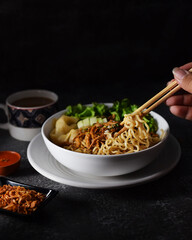 Mie ayam, noodles with chicken and vegetables in white bowl, Indonesian traditional food in dark and texture background. served with fried onions and a cup of coffee