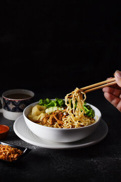 Mie ayam, noodles with chicken and vegetables in white bowl, Indonesian traditional food in dark and texture background. served with fried onions and a cup of coffee. hand holding chopsticks