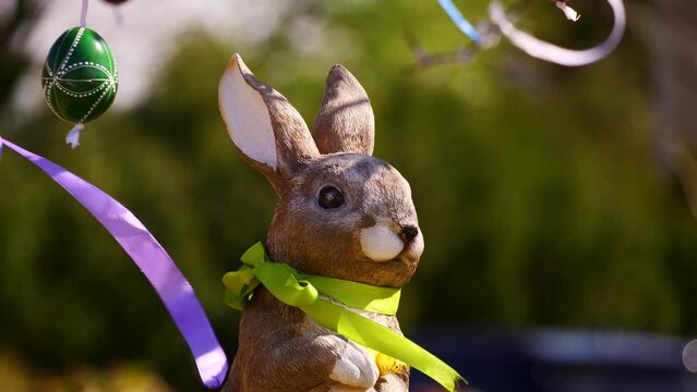 Easter in spring season with a cute rabbit decoration with a green scarf, and colorful blurred background includes trees, eggs , green grass.