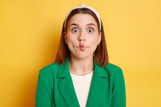 Funny optimistic woman wearing green jacket making fish lips grimacing having fun looking at camera with big eyes posing isolated over yellow background.