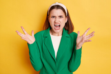 Angry irritated woman wearing green jacket posing isolated over yellow background arguing screaming...