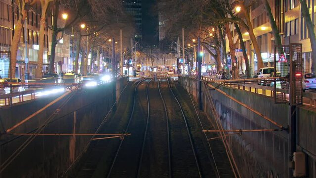 Timelapse with symmetrical arrangement of trams and traffic at the train station. Trams smoothly enter and exit while cars flow on the side. Tracks in the foreground provide a visual focal point.