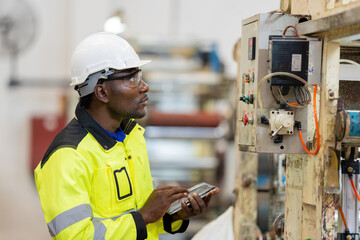 african engineer looking at the control panel Check the system via tablet. Wear uniforms and helmets. Around there are machines working on steel and plastic production in the industrial factory.