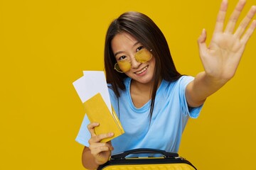 Asian woman traveling with yellow suitcase and tickets with passport in hand, tourist traveling by plane and train with luggage on yellow background in blue T-shirt and jeans