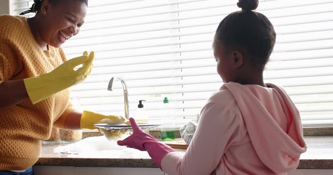 Happy african american mother and daughter washing up dishes in kitchen, slow motion