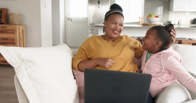 Happy african american mother and daughter relaxing on sofa using laptop, in slow motion