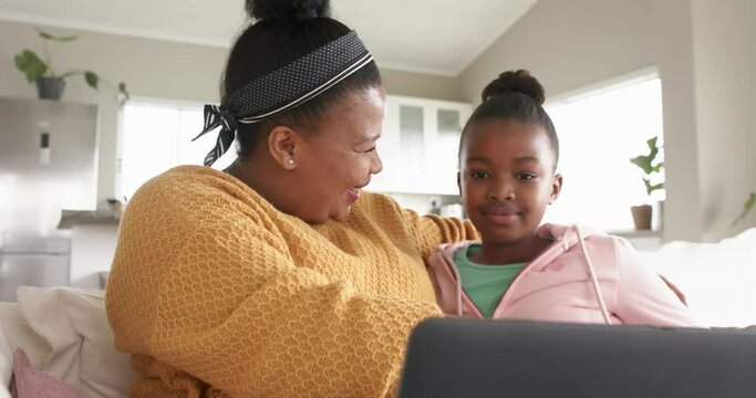 Happy african american mother and daughter relaxing on sofa using laptop and hugging, in slow motion