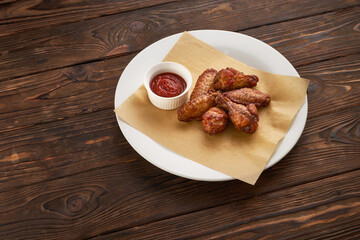 Grilled spicy crispy chicken legs and wings served with ketchup on a white plate over dark wooden rustic background, fast food and delivery concept for menu design